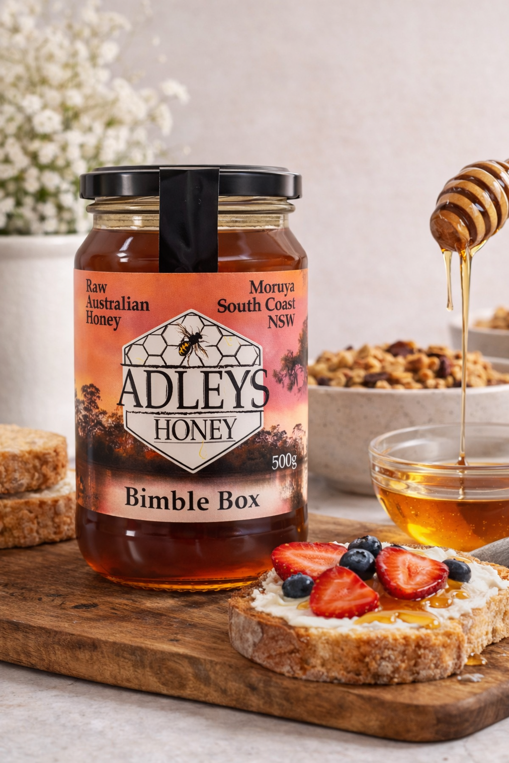 Adley's Honey jar with a bowl of honey, toast with honey and fruit, and a honey dipper on a wooden board.